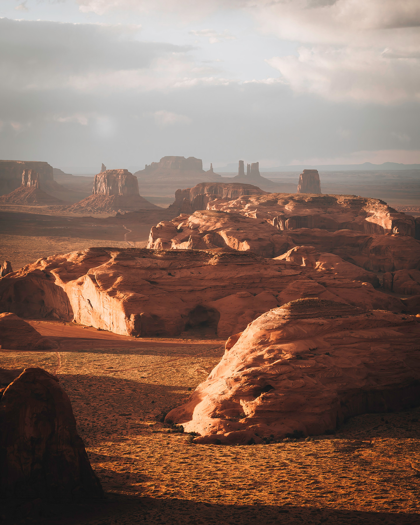 Monument Valley buttes and mesas from Hunts Mesa at golden hour — warm side-light raking across layered red-rock formations, hazy distant spires under a soft clouded sky.