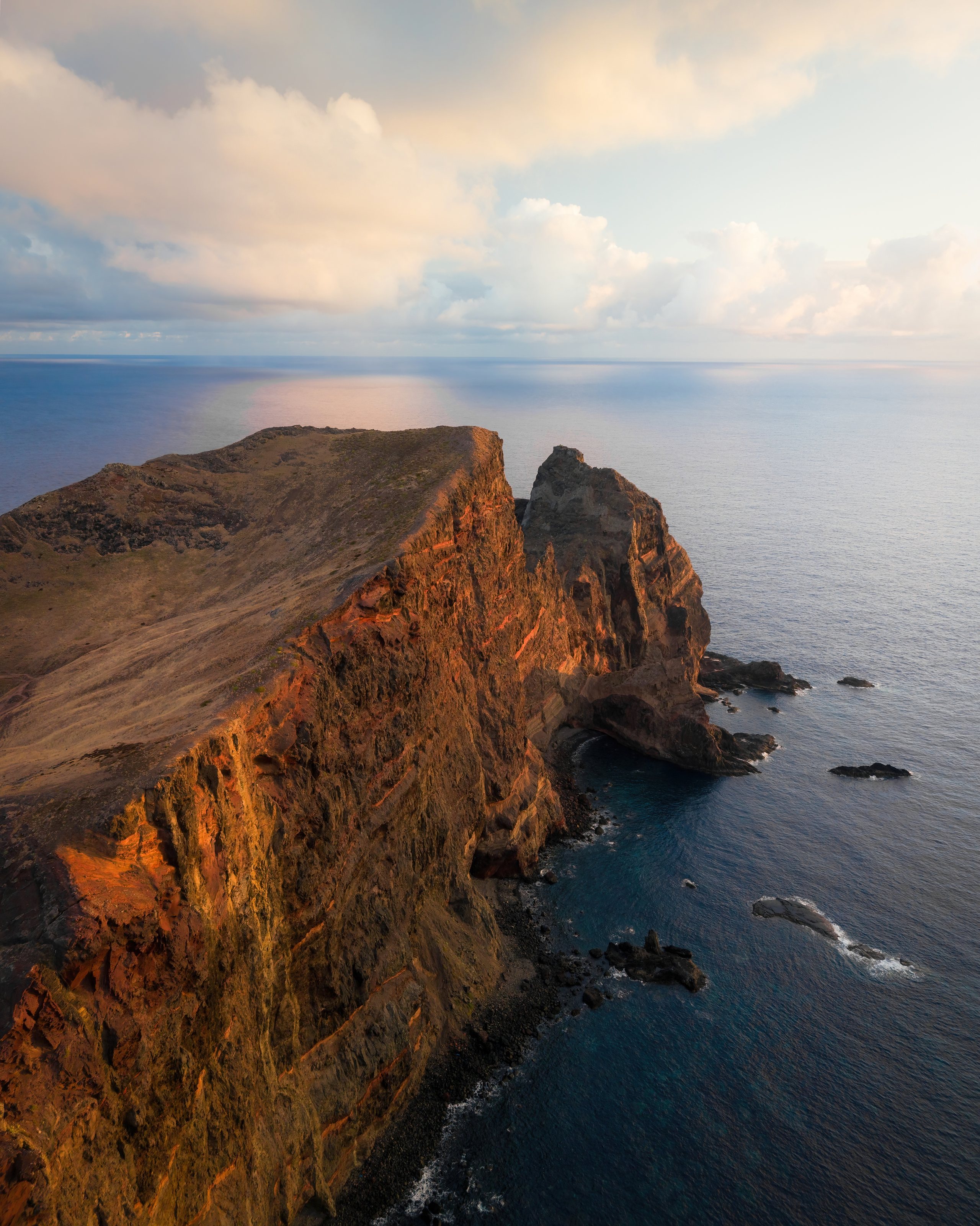 First light rakes across a volcanic headland above the Atlantic, the cliff face lit gold along its spine, deep blue water on both sides.