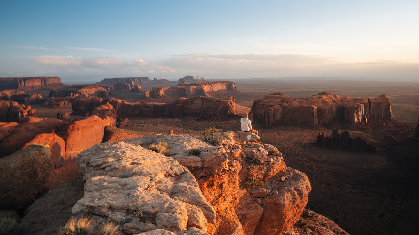 A lone figure sits on a red-rock ledge at sunrise above Monument Valley — sandstone buttes and mesas receding to the horizon in warm dawn alpenglow under a soft glowing sky, the vastness scaled by the small human form.