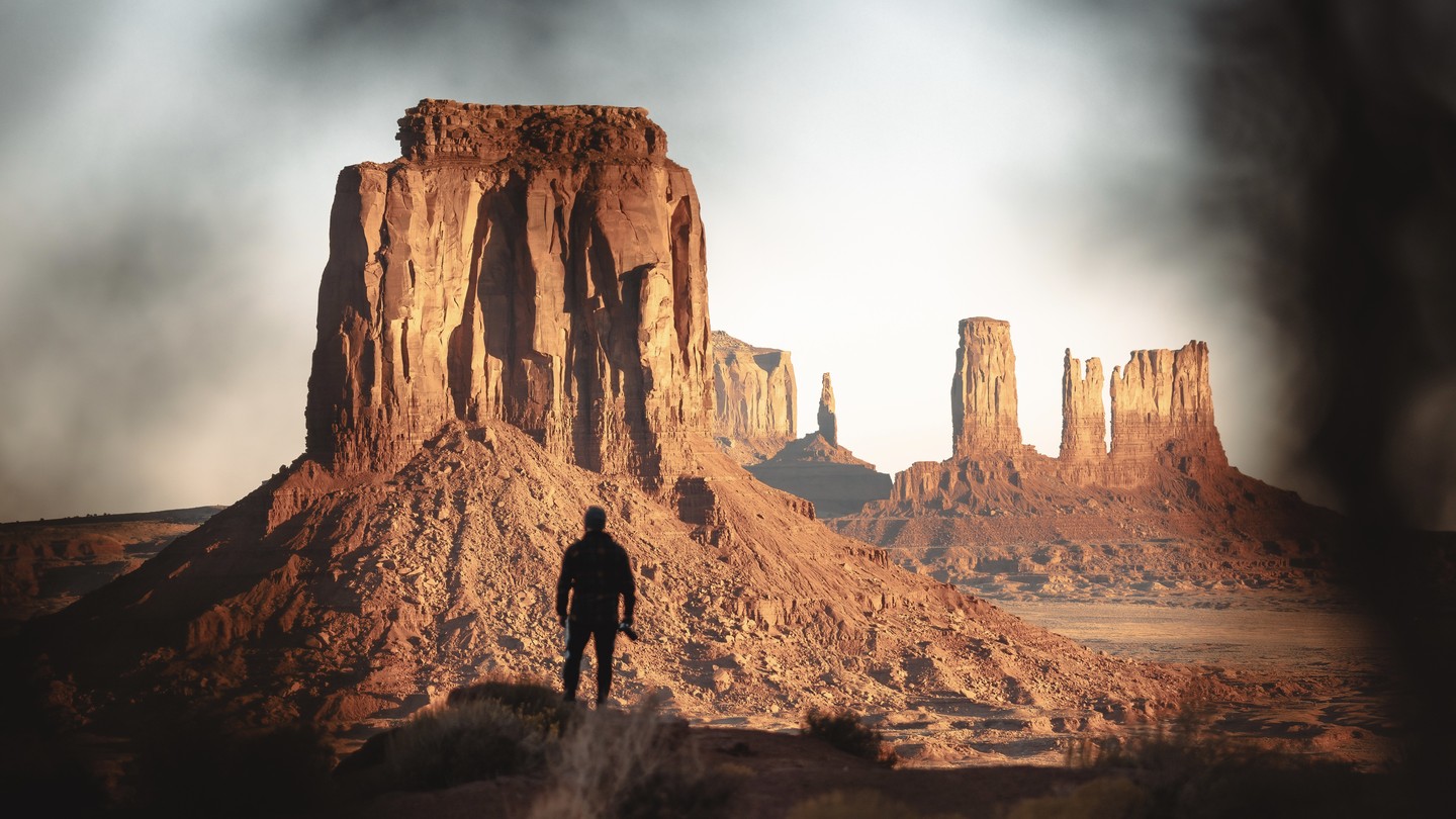 A figure standing at the base of a monolithic Utah desert formation at dawn — heroic-scale canyon walls in warm rim-light, the human form scaled by the geology, smaller monoliths in the distance, editorial light, the geometry of a guest-as-protagonist.