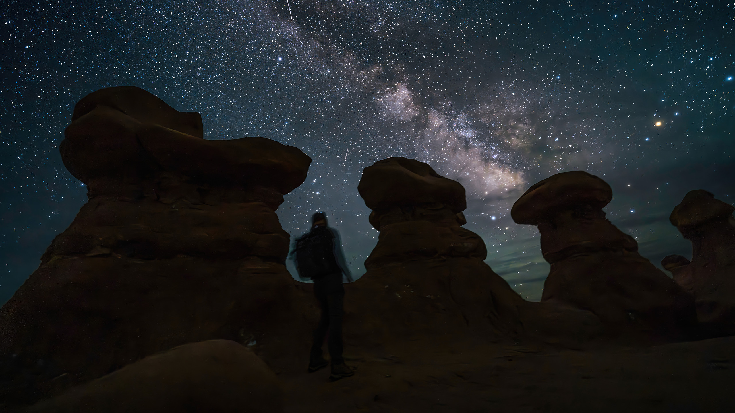 A lone figure stands beneath the Milky Way over the hoodoos of Goblin Valley at night — the galactic core arcing across a star-dense sky, the dark desert scaled by a single silhouette.